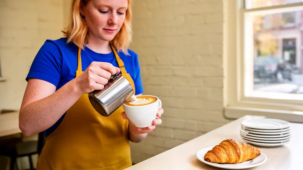 A café owner in a mustard apron pouring steamed milk into a white cup to make latte art at a sunny counter.